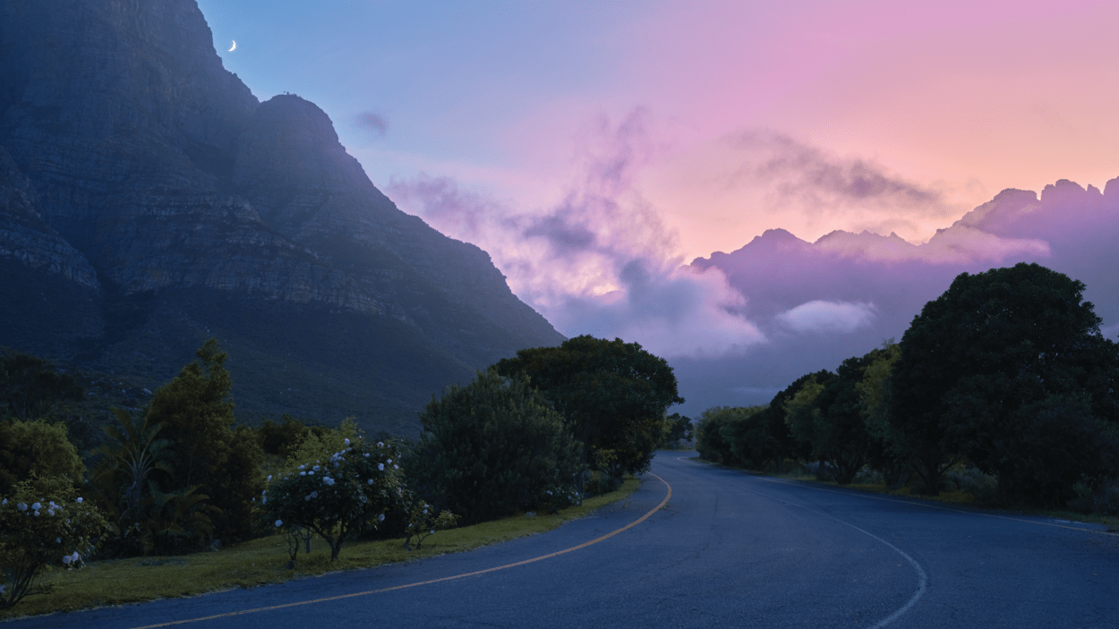 Tom vei med trær på hver side, fjell og rosa himmel i bakgrunnen.